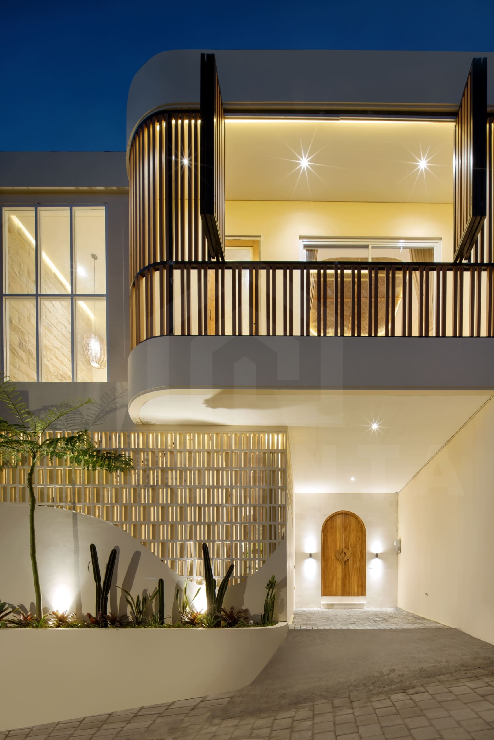 Modern villa facade with curved balcony, warm lighting, wood accents, and landscaped entrance in Bali
