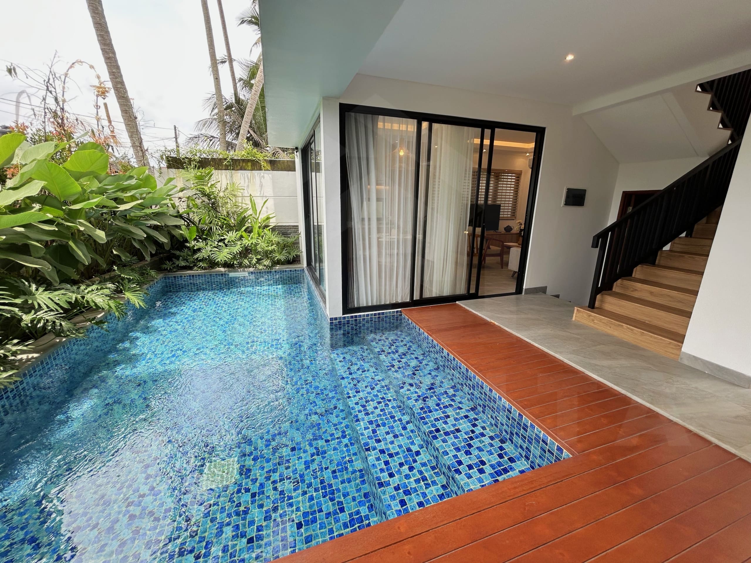 Compact villa pool with blue mosaic tiles beside bedroom sliding glass doors and lush plants.