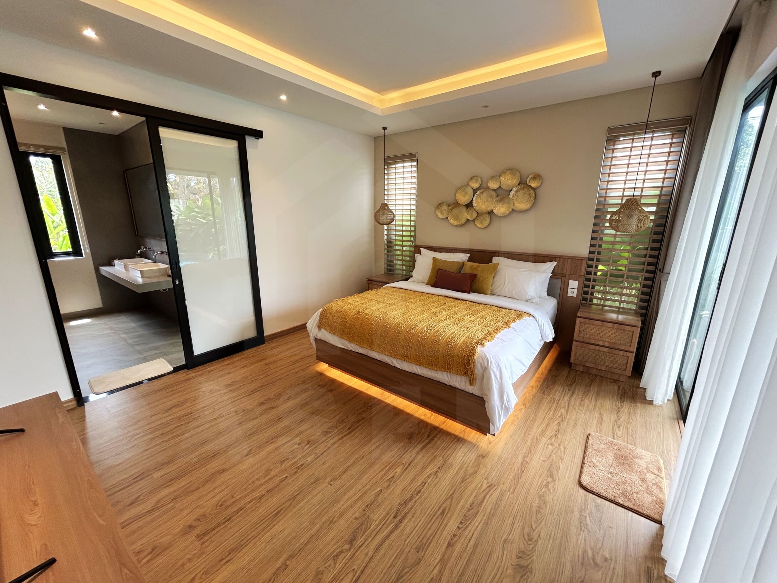 Modern master bedroom with warm lighting, wood flooring, and sliding door to ensuite bathroom.