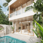 Modern two-story glass-walled home surrounded by lush tropical plants and a rectangular swimming pool in the foreground.